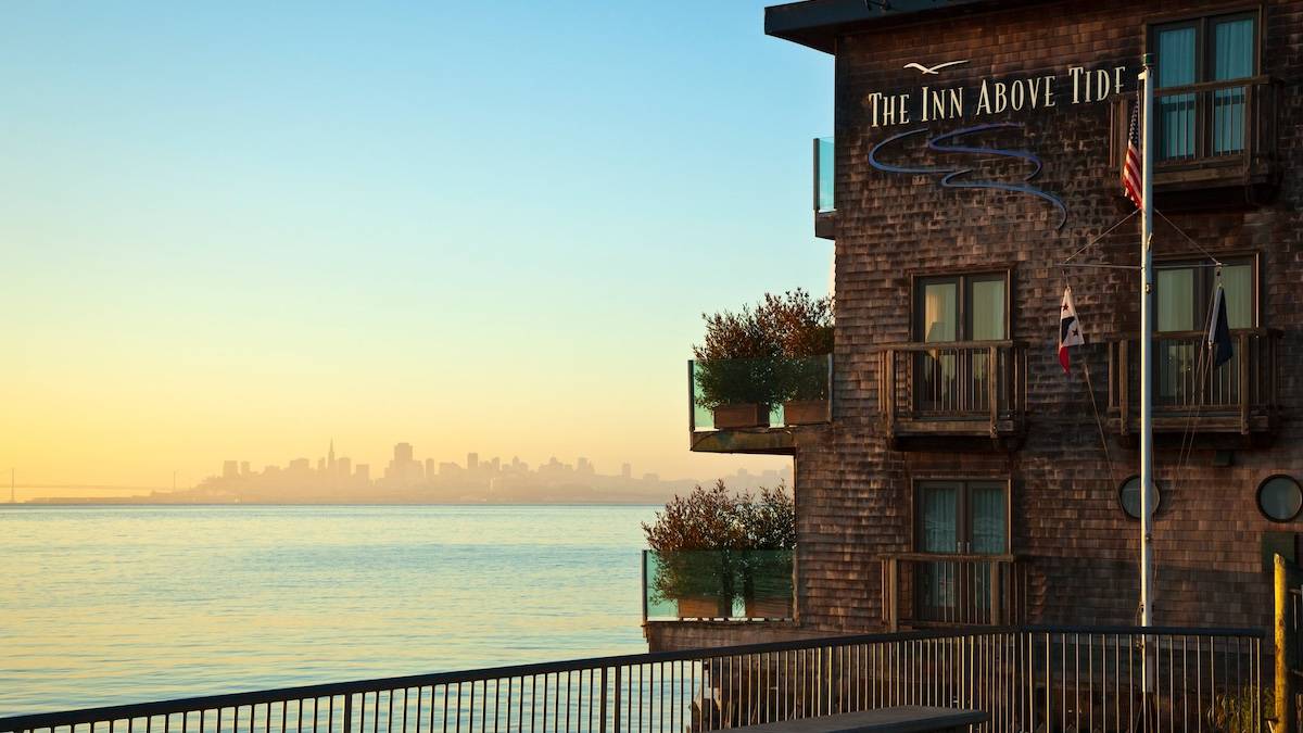 Side of a wooden shingled building with glass enclosed patios next to a bay with San Francisco in the background