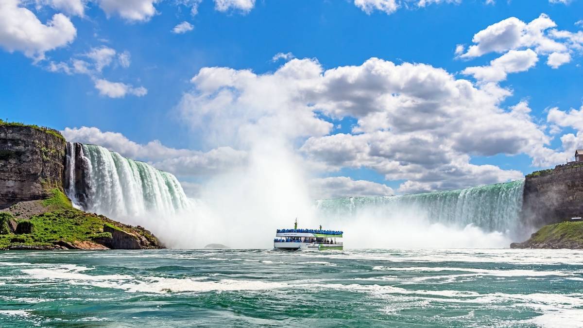 Ferry boat in water under a blue cloud studded sky with niagara falls in the background
