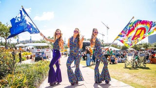 Three women in 70s disco clothing standing on a walkway at a festival holding flags
