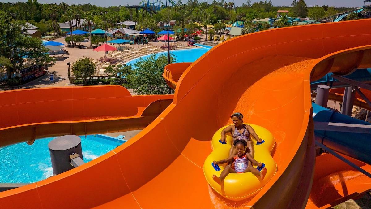 Orange water slides with two women in a yellow tube sliding down with a water park in the background