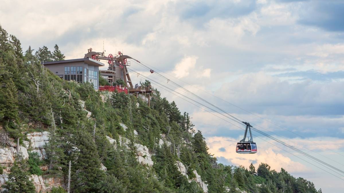 Aerial tram moving down cables over a wooded mountain under a cloudy blue sky