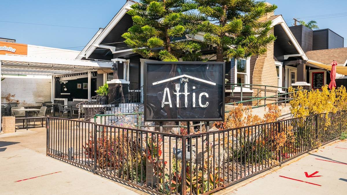 Black and white sign reading "The Attic" in a fenced in area in front of trees and a house that's been turned into a restaurant