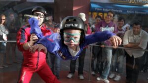 Kid indoor skydiving with an instructor behind him holding onto his legs as a crowd watches from the background