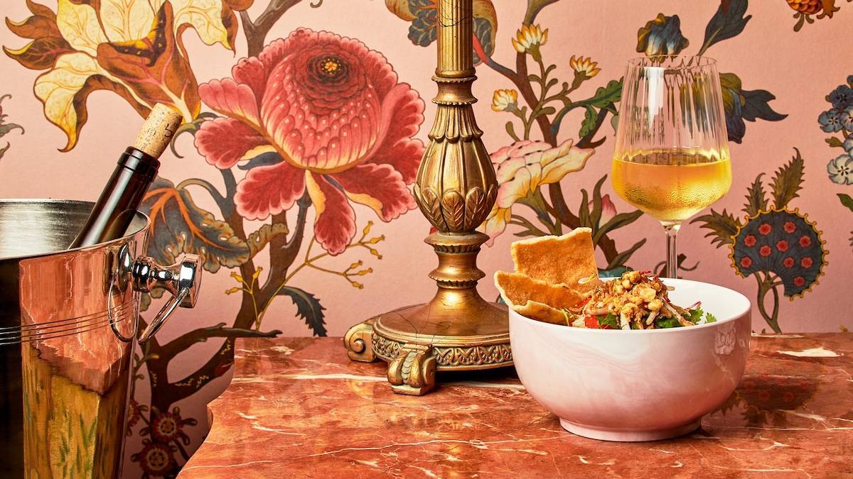 Glass of white wine next to a white bowl full of fresh food with pita chips on a table in front of pink and floral wall paper with a wine chiller and lamp base nearby