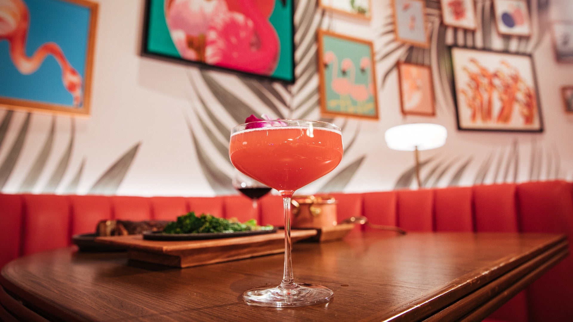 Pink cocktail sitting on a table with tropical wallpaper and flamingo art on the walls in the background