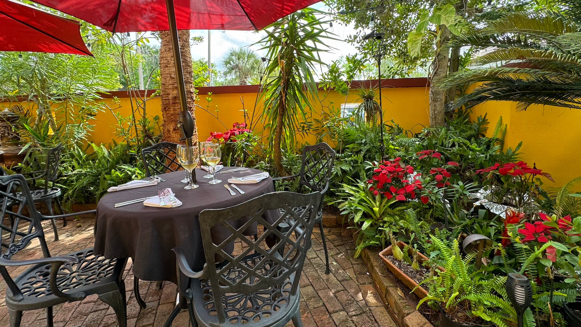 Outdoor space with yellow stone walls and a cobblestone floor with black patio furniture and red umbrellas surrounded by plants