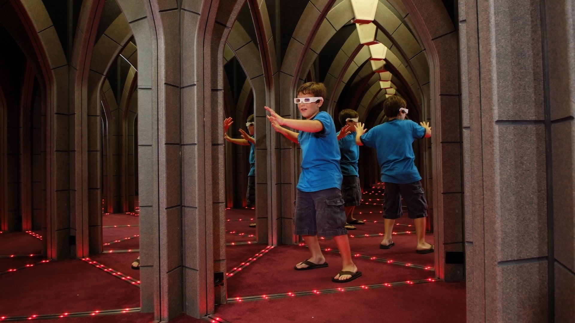 Boy in a blue shirt and 3D glasses on in a mirror room with his arms stretched out in front of him