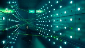 Person crouching down in a room with green lasers and metal walls with lights studding the walls