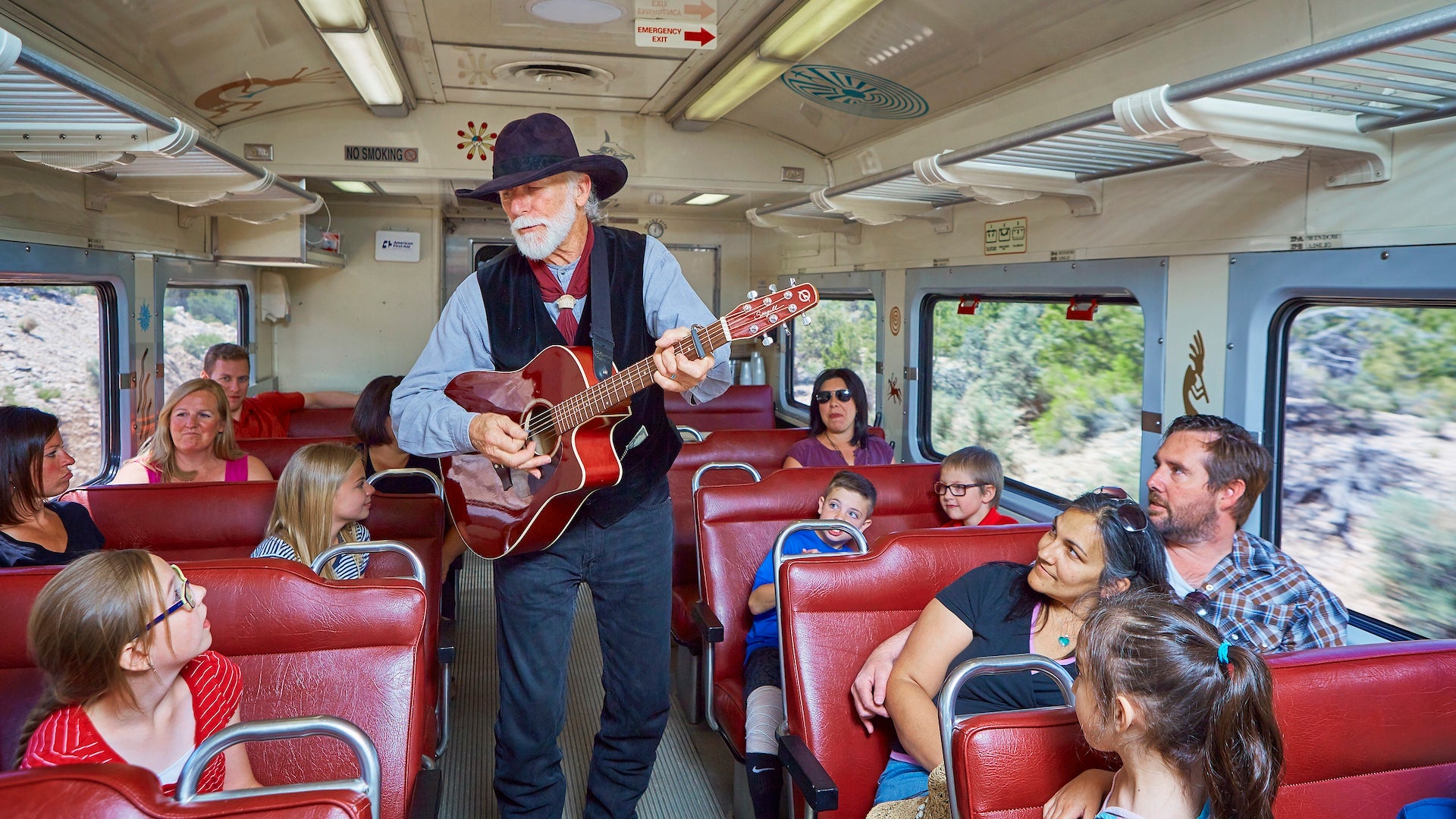 Man in western wear playing a guitar to a train car full of people sitting in red booths