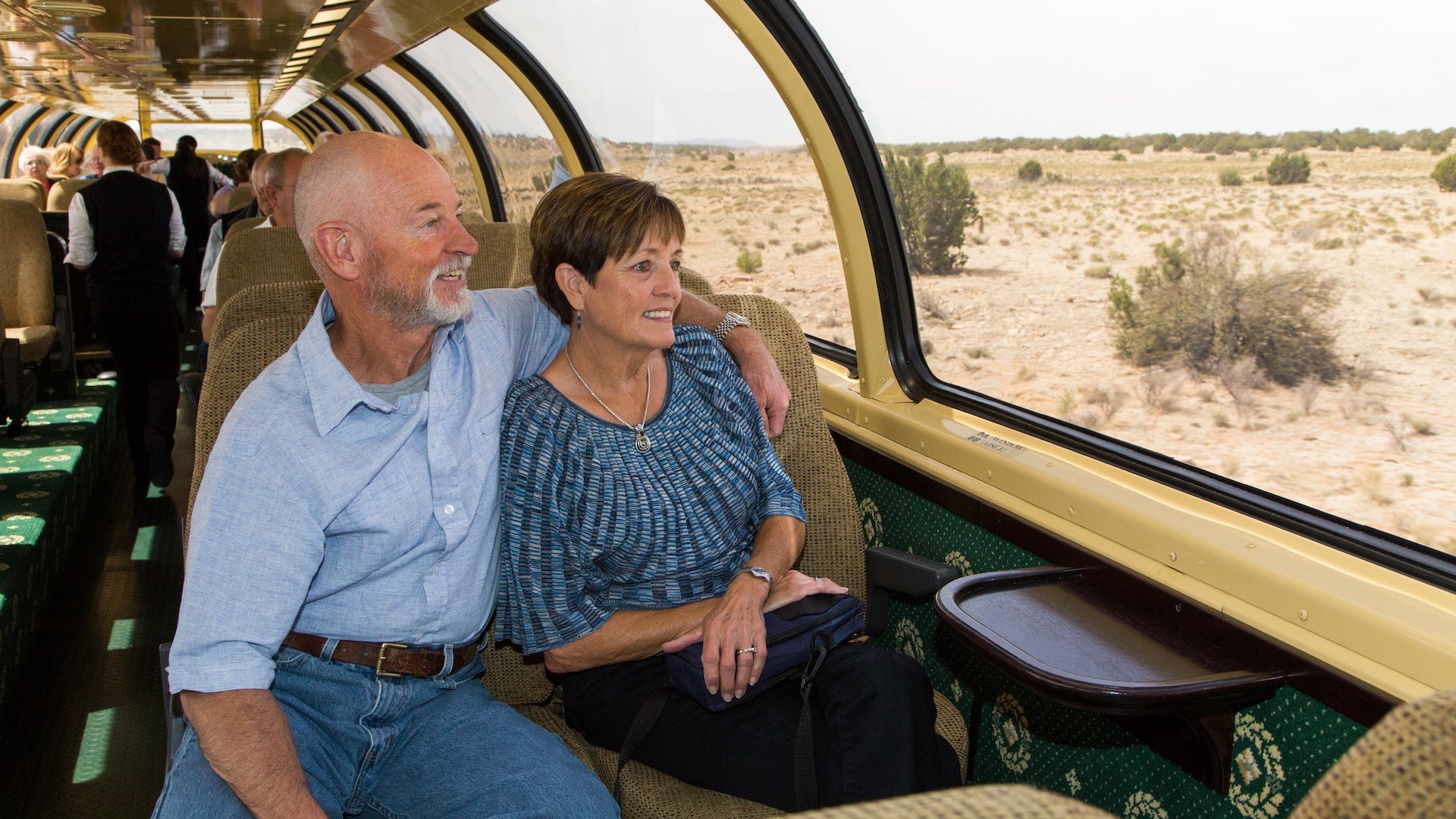 Interior view of a luxury train car with an older couple sitting on a booth looking out of large windows at the desert.
