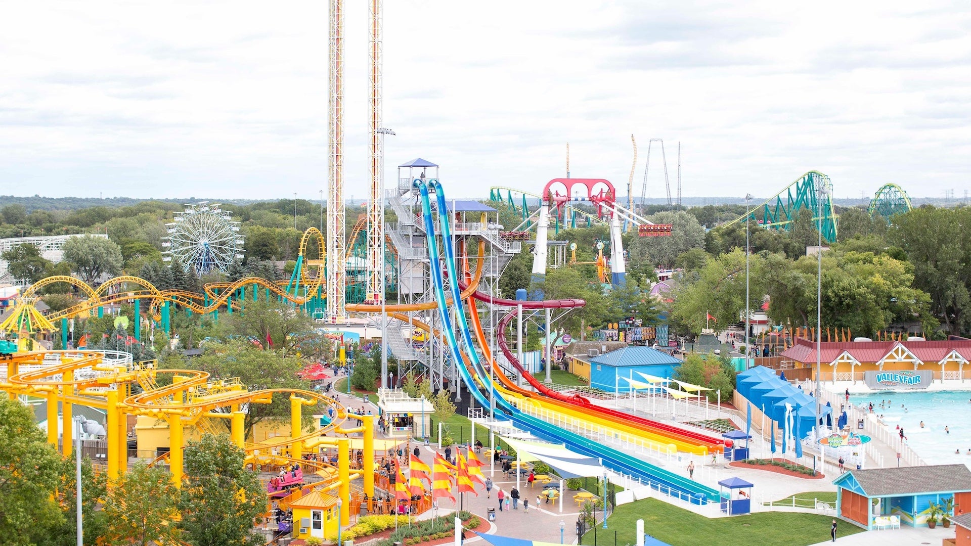 Wide shot of a theme park with multiple roller coasters, a ferris wheel, water park and slides all surrounded by green trees.