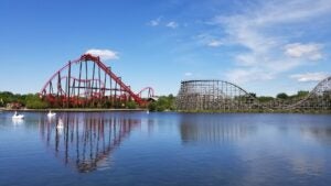 Wide shot of two roller coasters next to a lake under a blue sky.