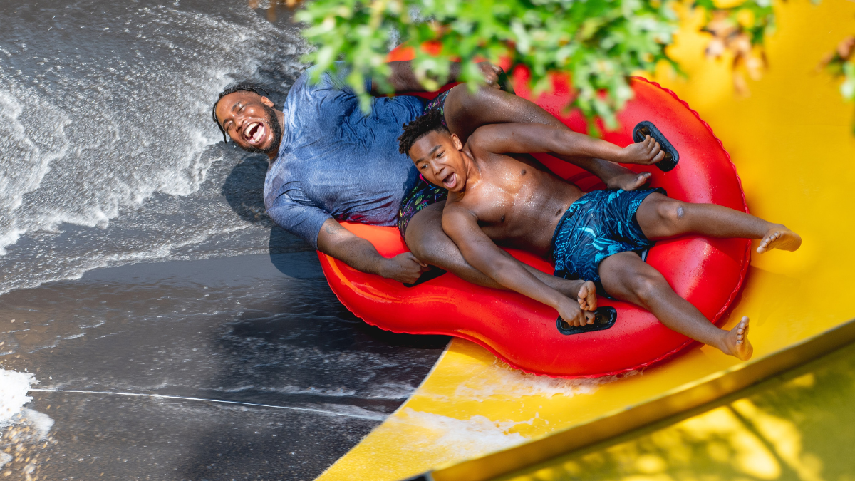 A father and son laughing together as they ride a red inflatable down a water slide.