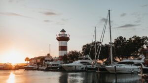 Red and white striped lighthouse next to a marina with boats at sunset