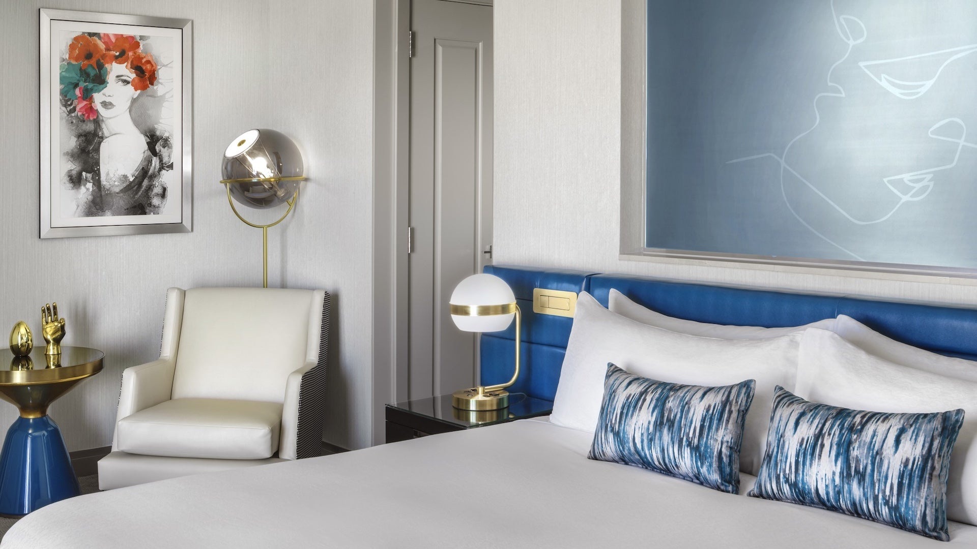 White bed with blue pillows in a white walled room with abstract art and a bedside table with a lamp