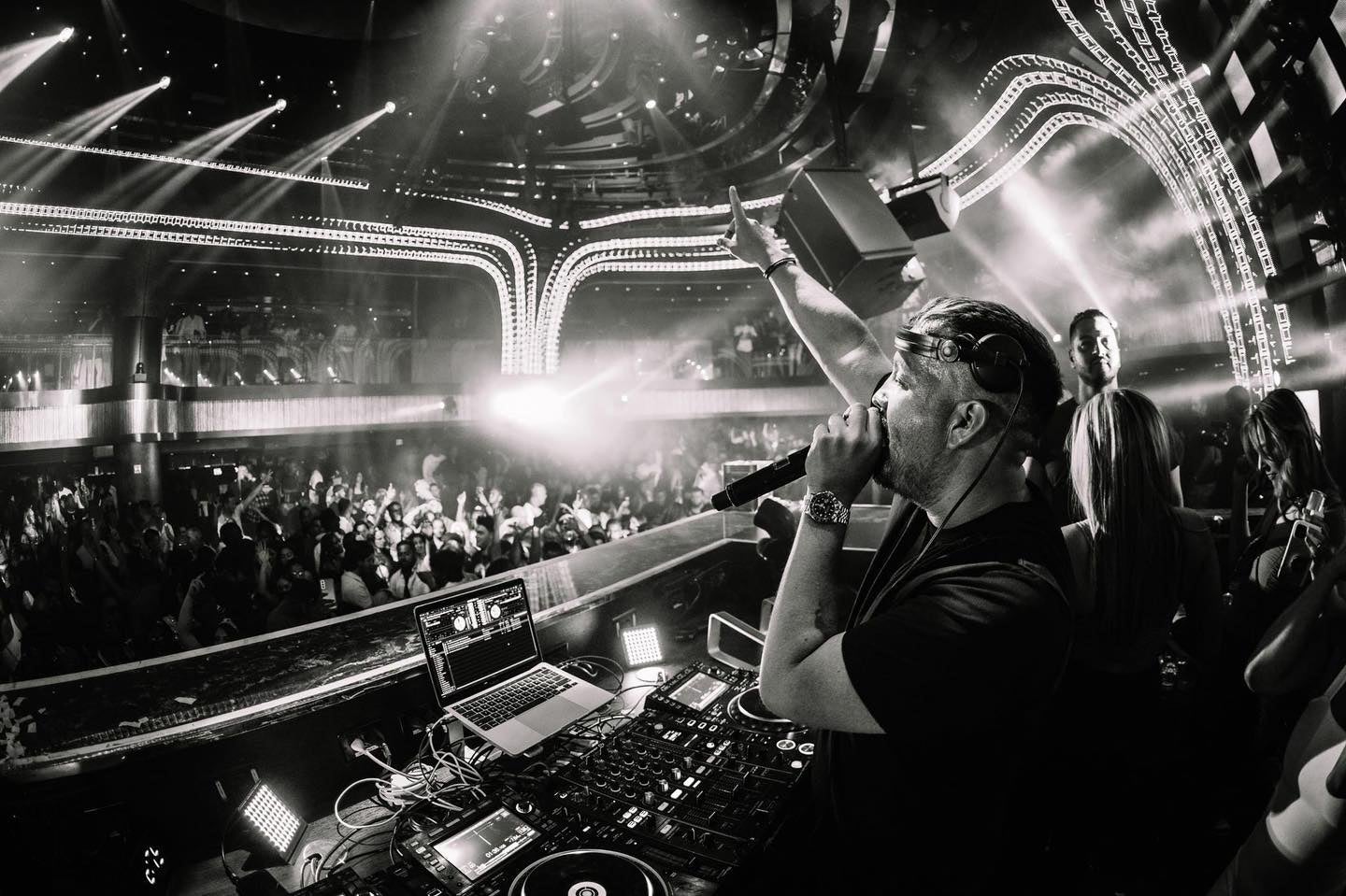 Dimly lit night club with people dancing in the crowd and a man standing on a stage with a microphone and one arm pointing to the ceiling