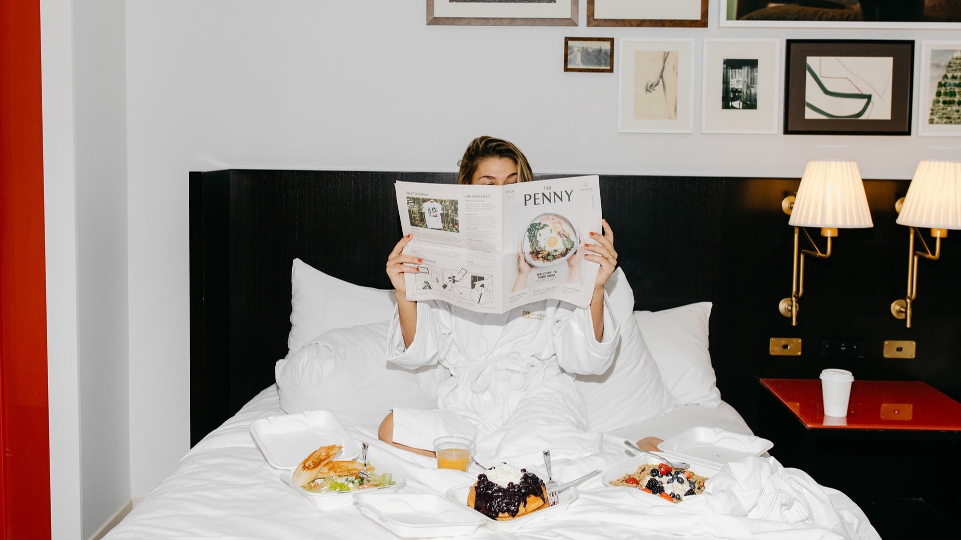 woman reading a newspaper in bed with food spread out, hotel room