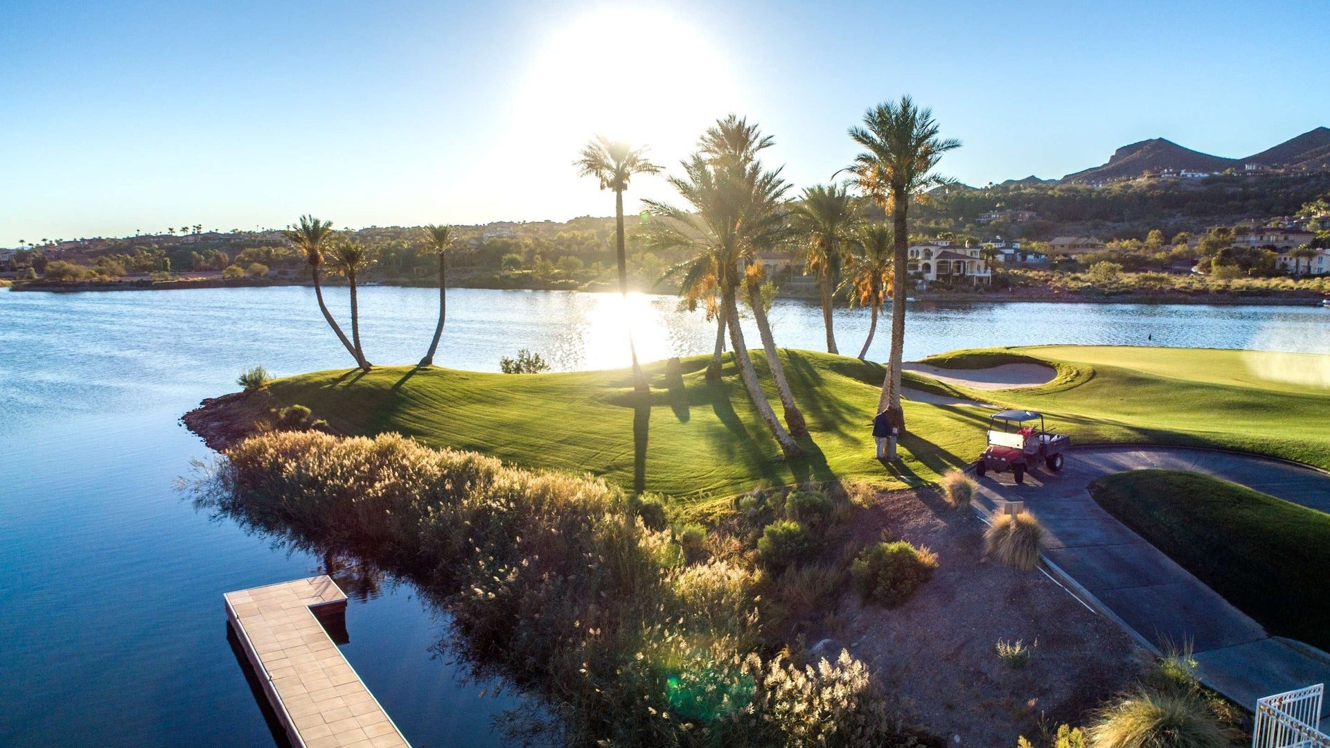 golfing greens beside a lake, palm trees and golf cart with a view of mountains in the back