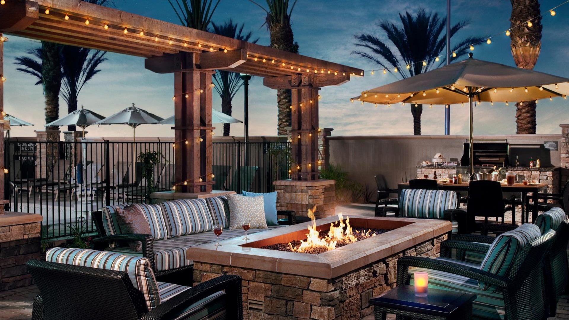 firepit surrounded by outdoor couches and chairs, barbecue grill on the side and outdoor umbrellas with lights