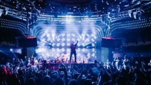 Dimly lit night club with people dancing in the crowd and a man standing on a stage with a microphone and one arm pointing to the ceiling