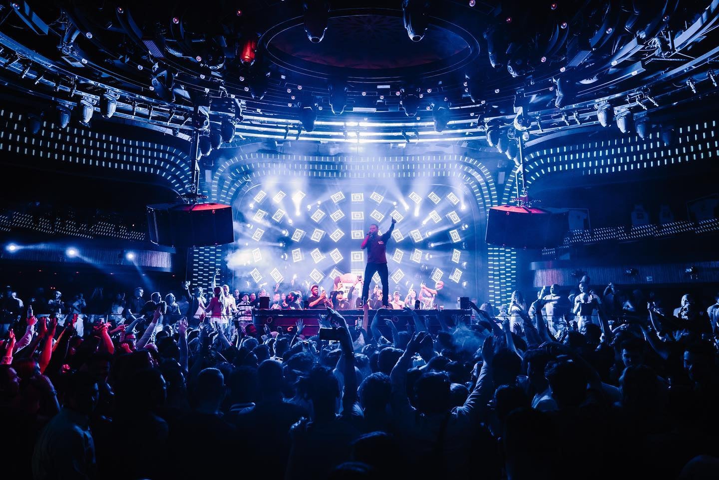 Dimly lit night club with people dancing in the crowd and a man standing on a stage with a microphone and one arm pointing to the ceiling