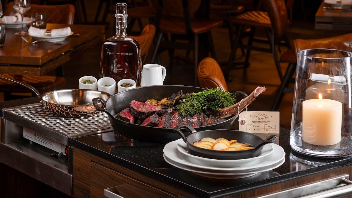 A cast iron skillet with meat in it on a table with bourbon and a frying pan and another pan full of potatoes