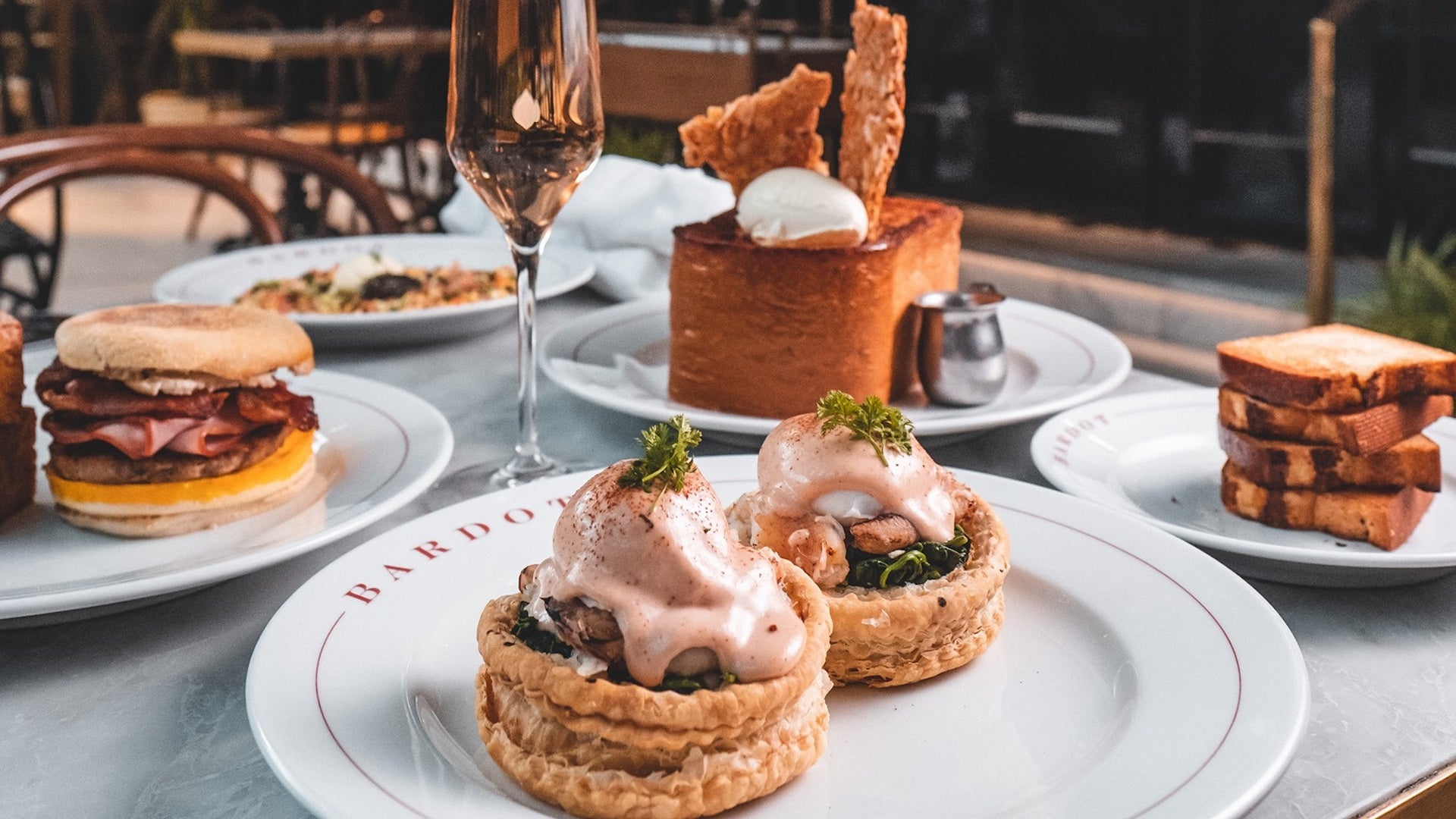 Several white plates laden with breakfast food on a table with a single glass of champagne