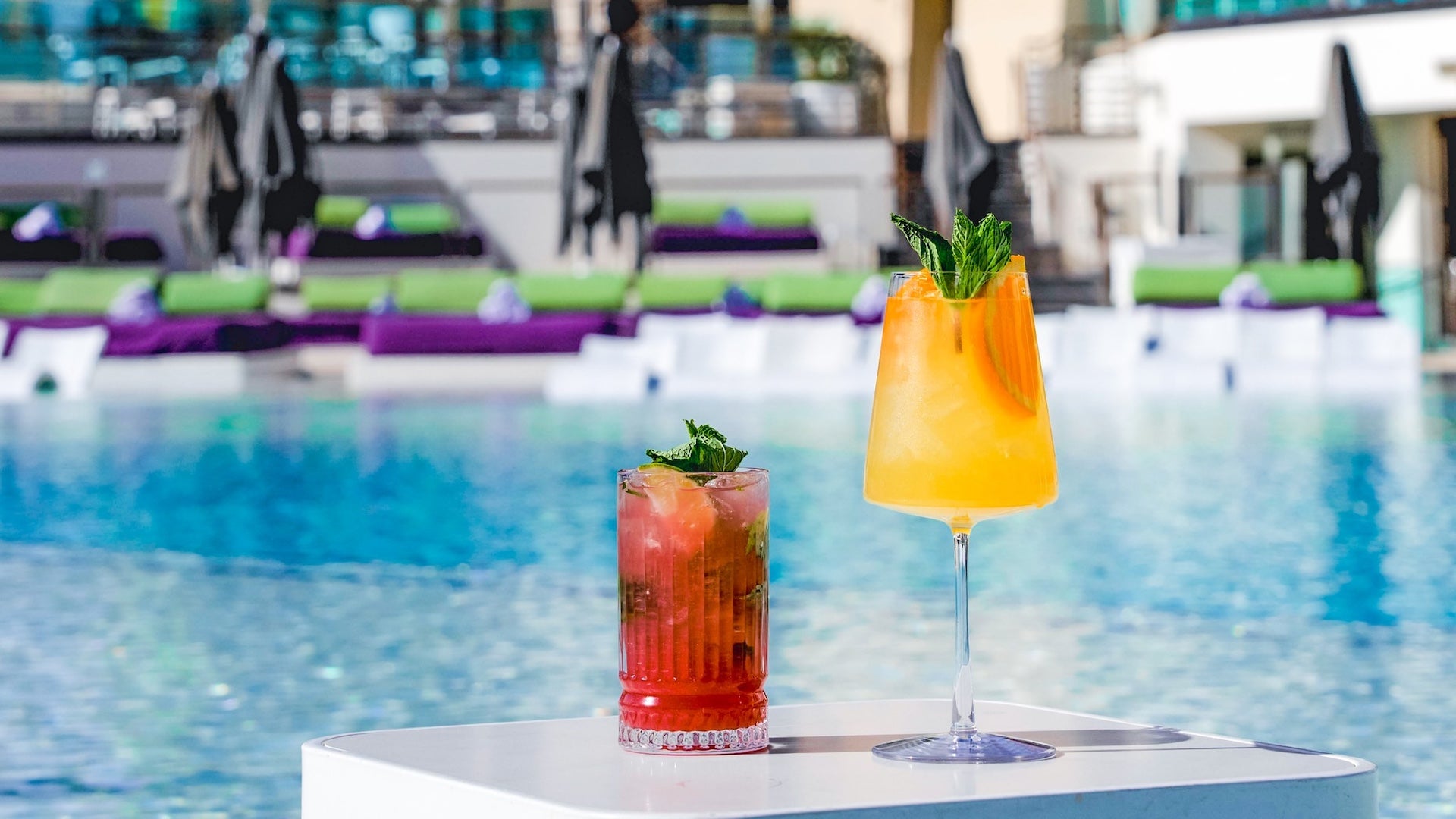 Two fruity cocktails on a white table with a pool in the background