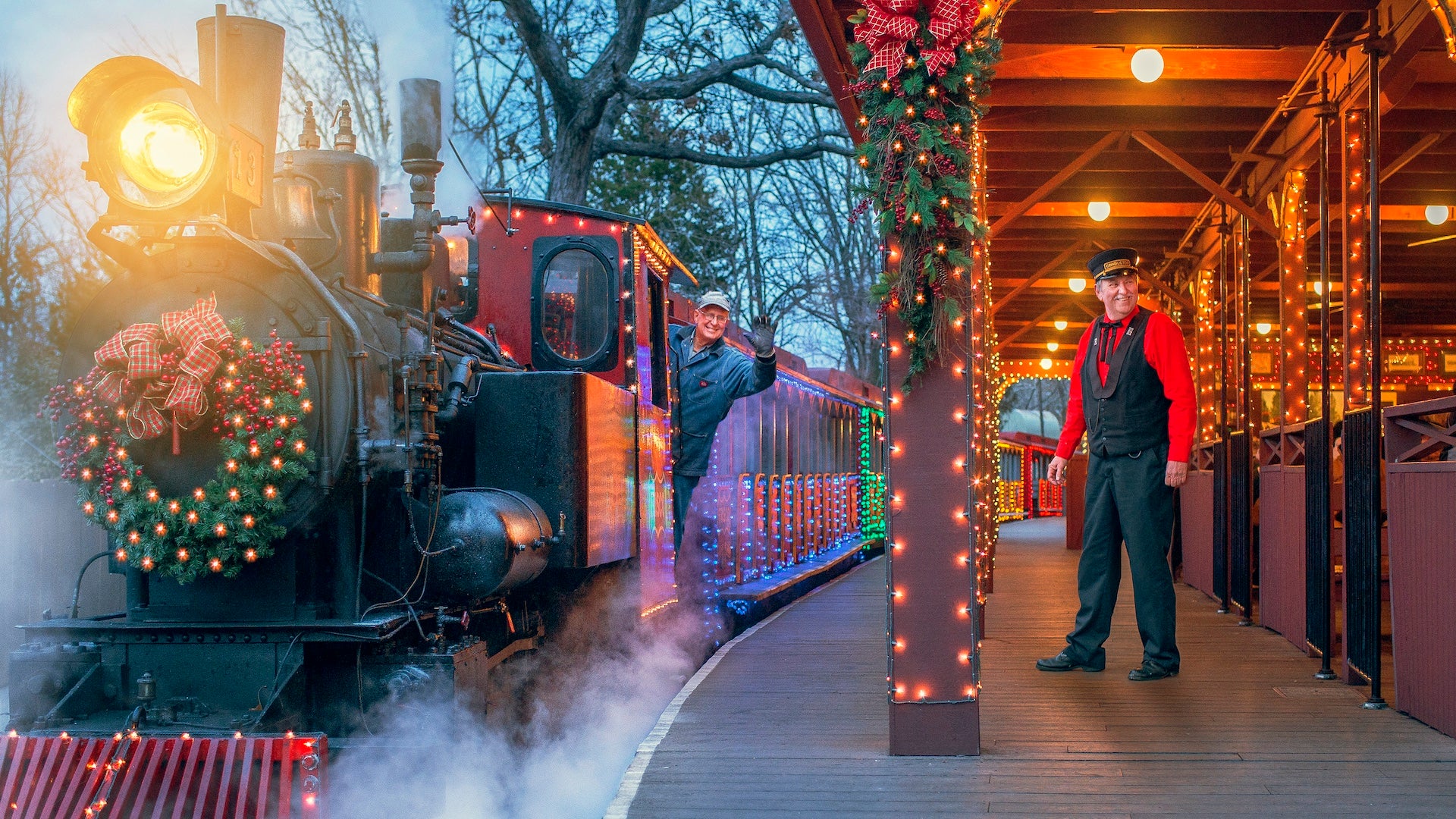 A conductor hanging from the side of a train with christmas lights on it next to a train depot with another man standing nearby