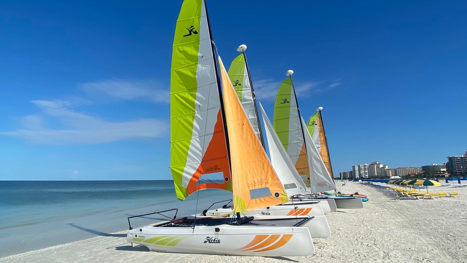 boats with sails on a shore with beach cabanas at the back