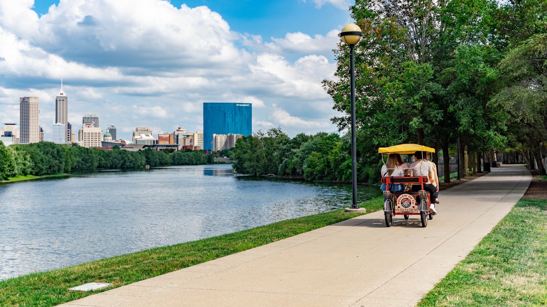 people riding a vehicle beside a river, skyline at the back and trees on the side