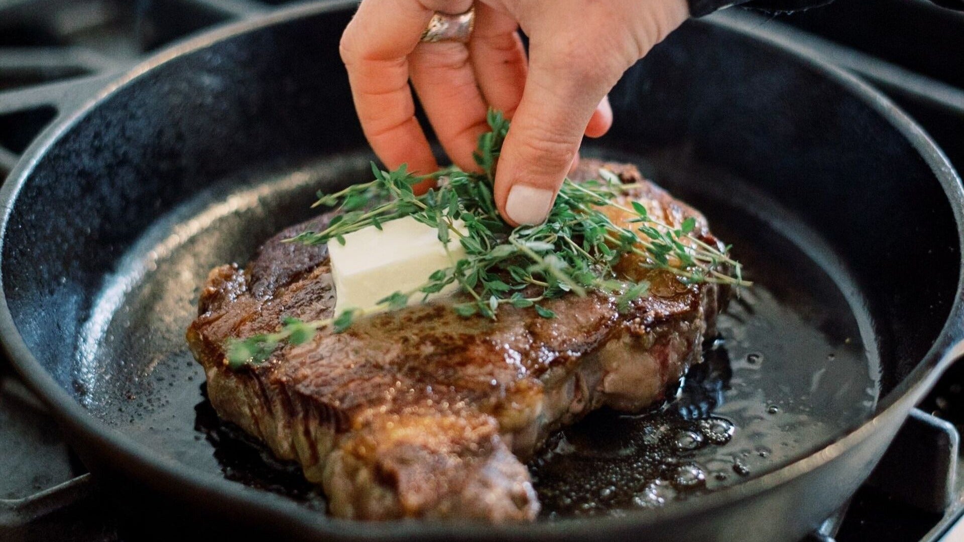 woman cooking steak, adding thyme and butter in a pan