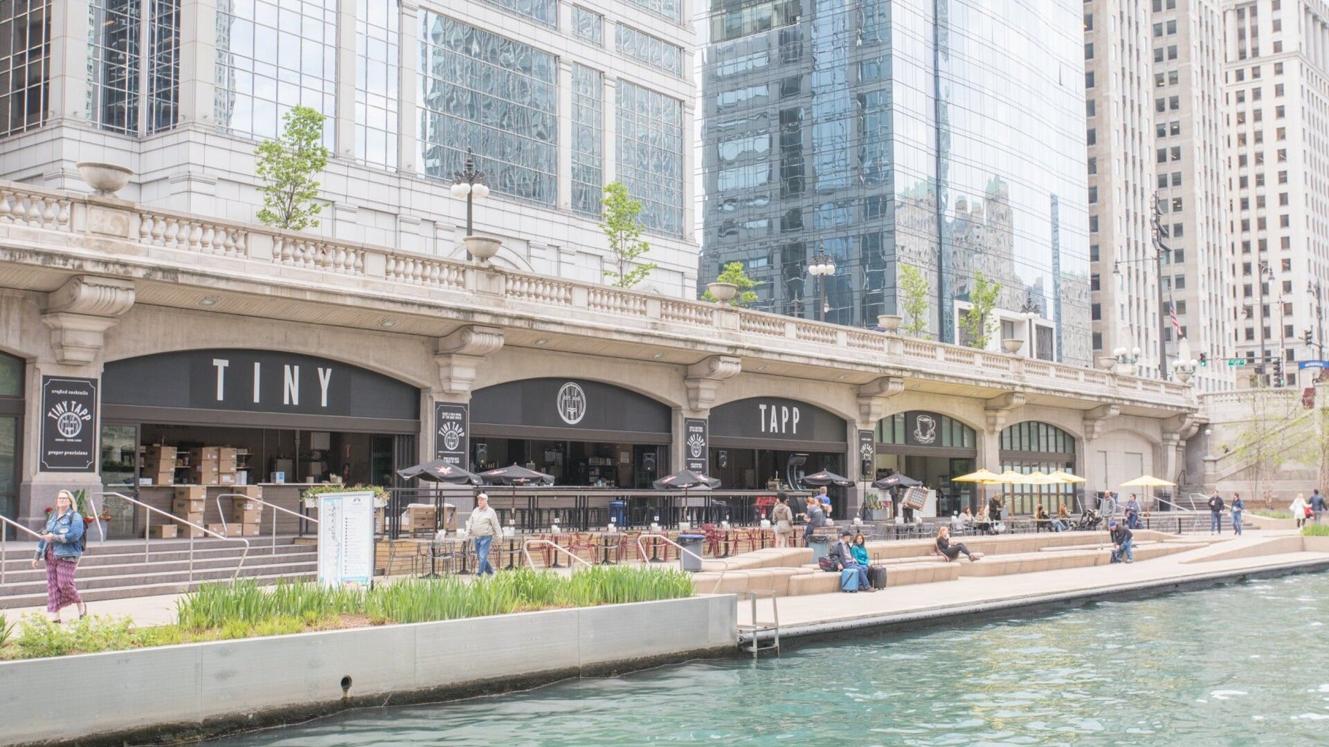 people sitting along a riverwalk with restaurants at the back