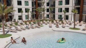 people in a pool with poolside chairs, palm trees, and hotel at the back