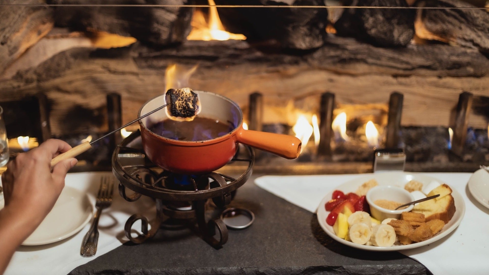 smores and fondue, with a plate of fruits on the side and a fireplace