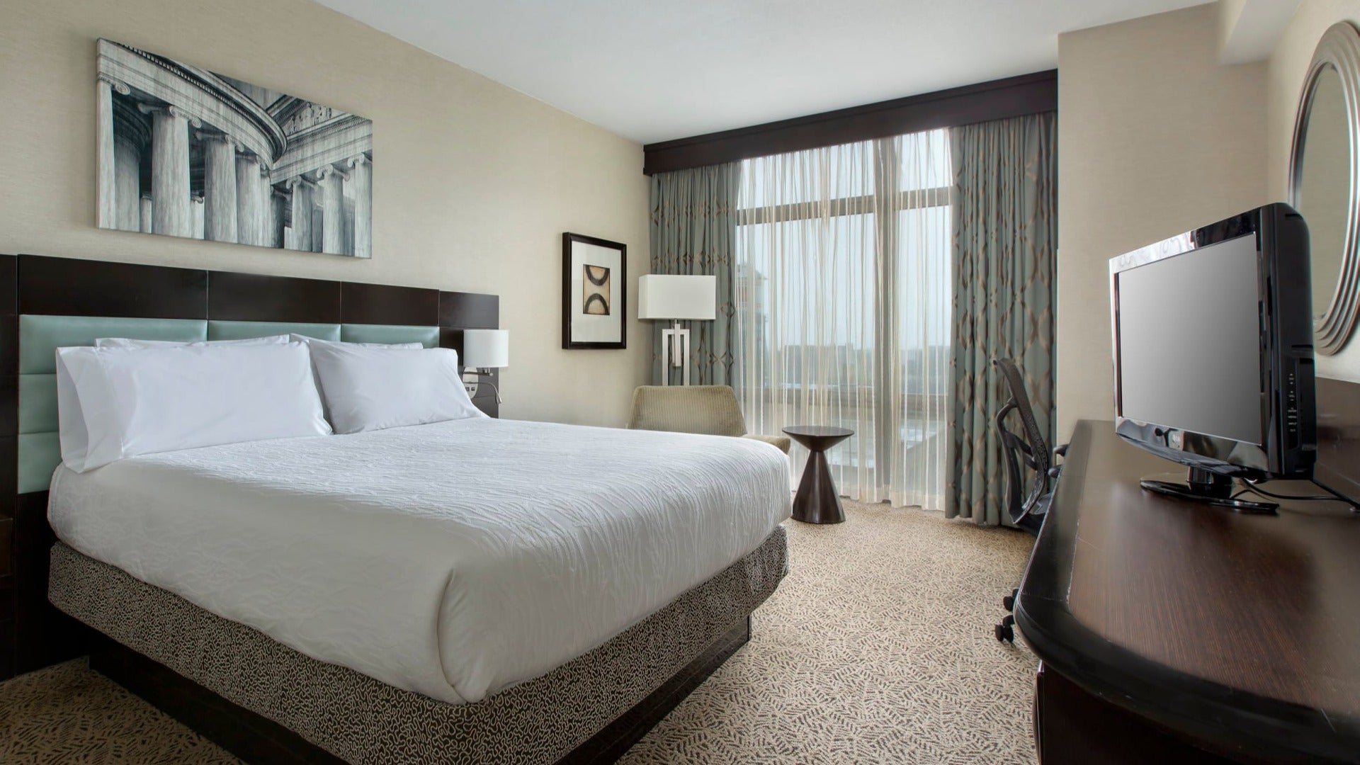 hotel room with bed, tv, lamp, painting, and curtains