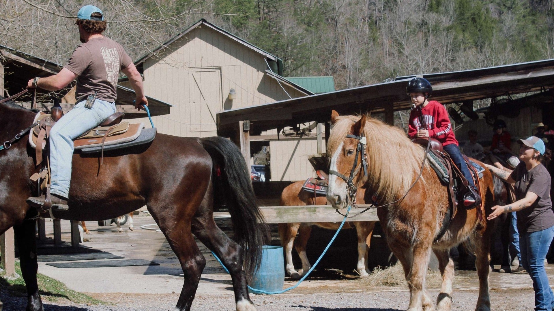 child riding a horse with a guide helping him, stables at the back