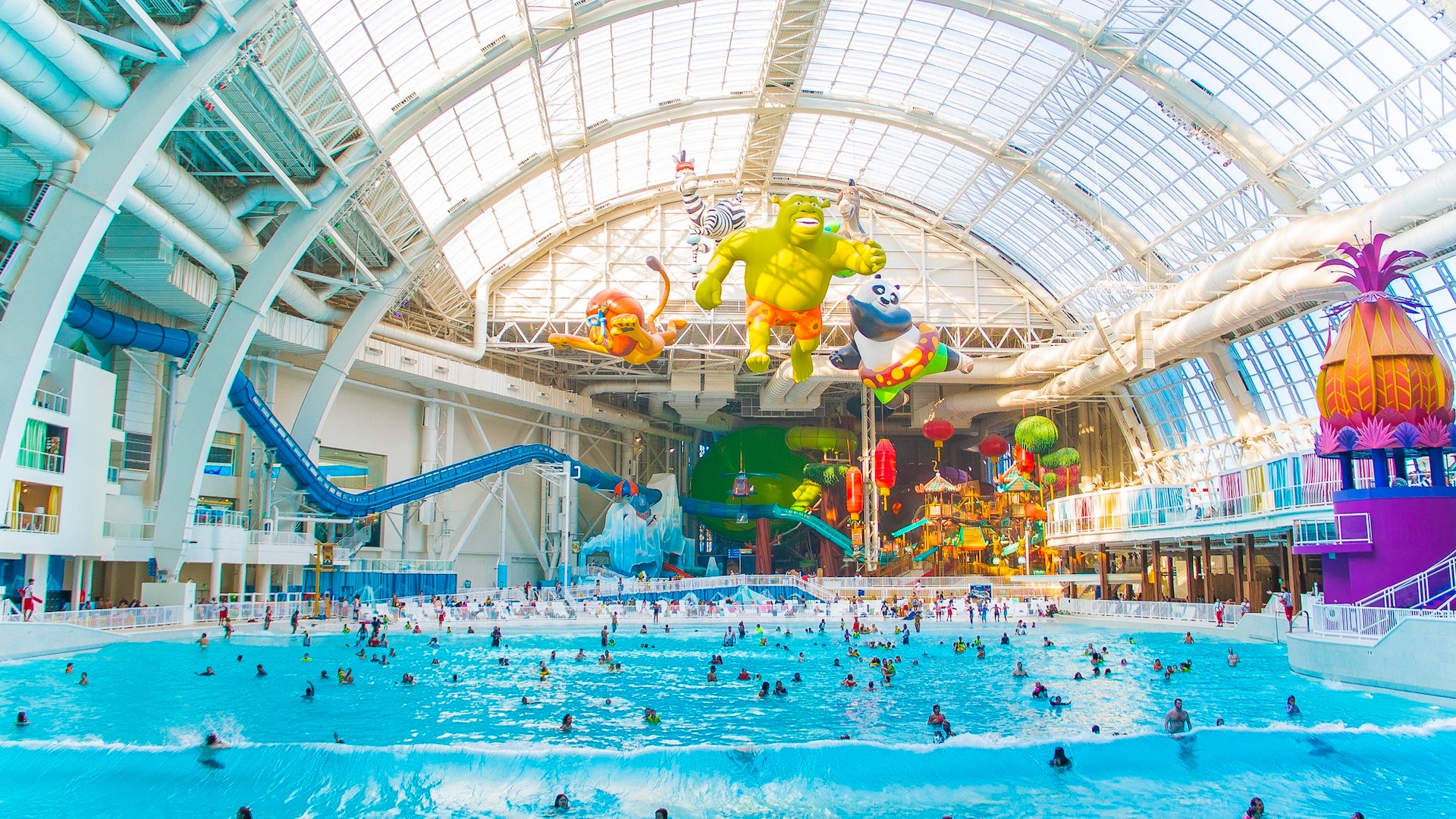 A large wave pool at an indoor water park filled with people swimming and a large water slide apparatus in the background