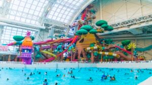 A large wave pool at an indoor water park filled with people swimming and a large water slide apparatus in the background