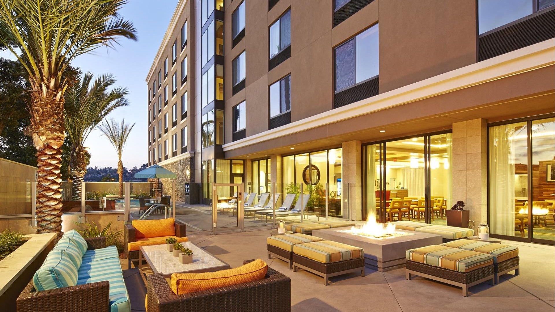 hotel lounge area with fire pit, sofa, chairs, palm trees and pool at the back