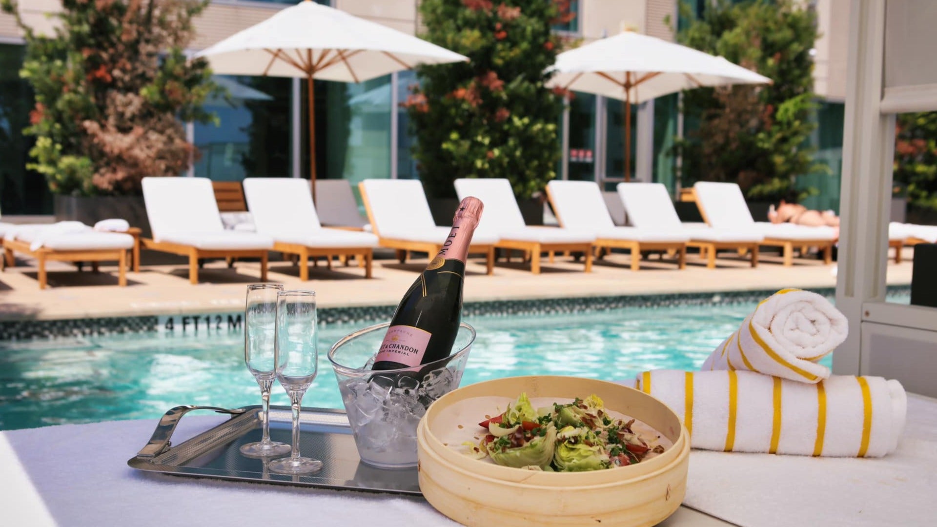 bottle of wine with wine glasses, salad, towels, pool chairs and umbrellas at the back