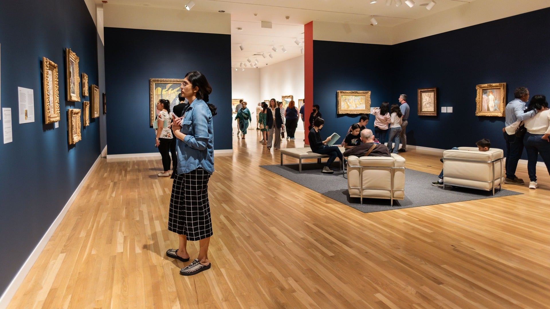 people admiring artworks in a room, paintings hanging on the walls with a seating area in the middle