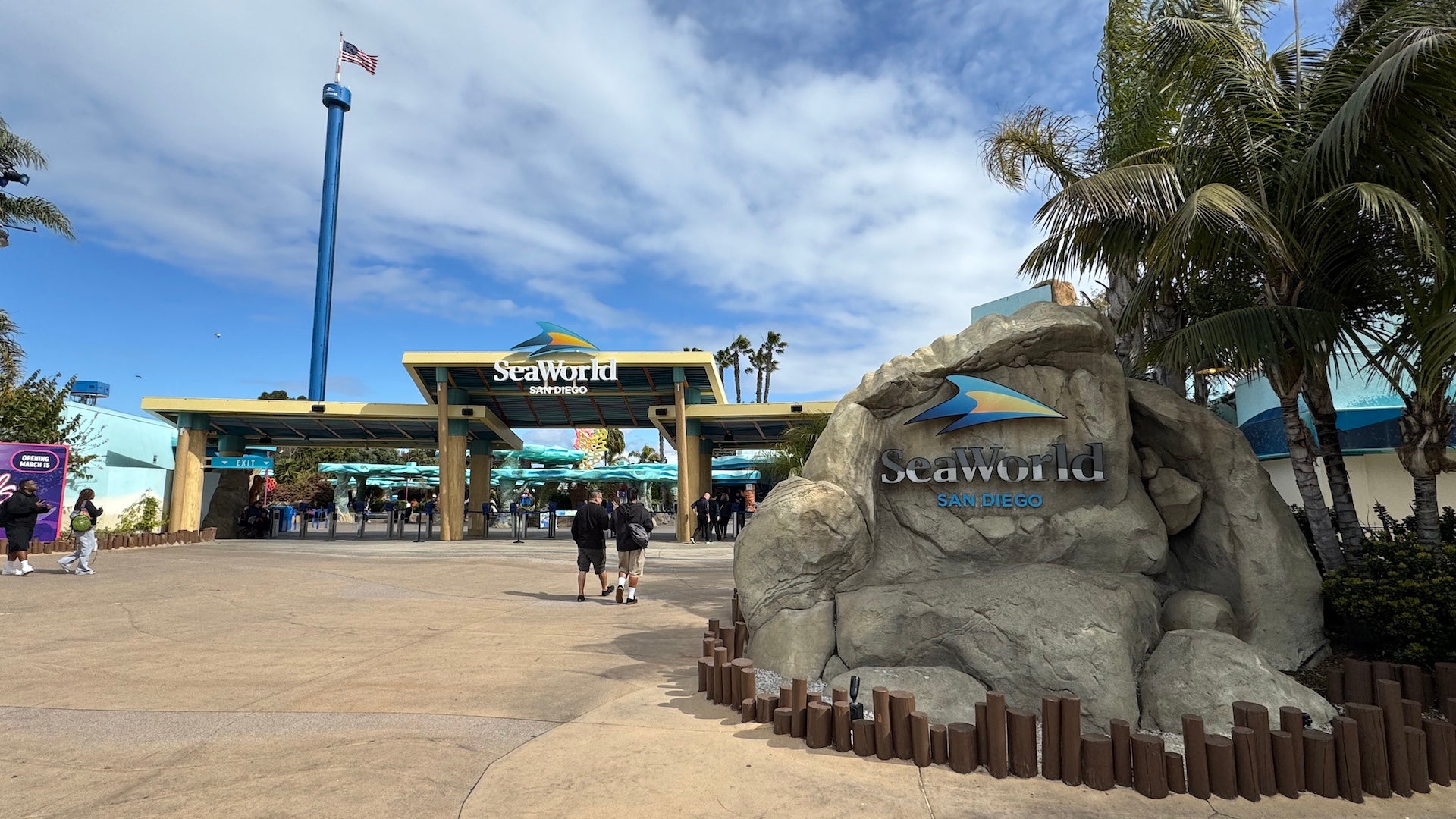 Entrance to SeaWorld San Diego with a large stone sign, palm trees, and guests walking toward the gates.