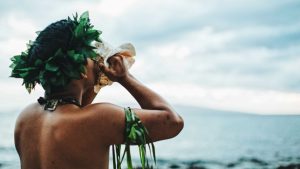 man in a luau attire blowing through a conch shell
