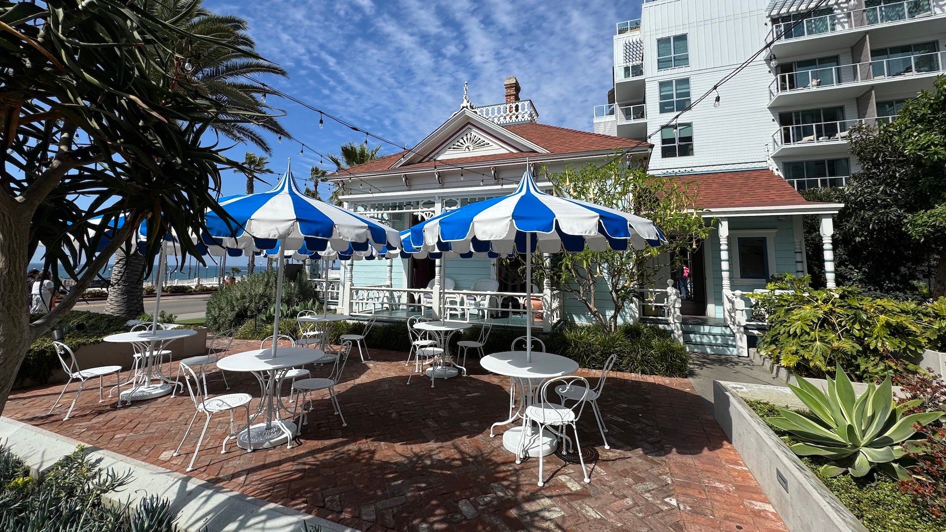 Brick patio outside the Top Gun House with white café tables, blue-and-white striped umbrellas, and views of the ocean and palm trees in the background.
