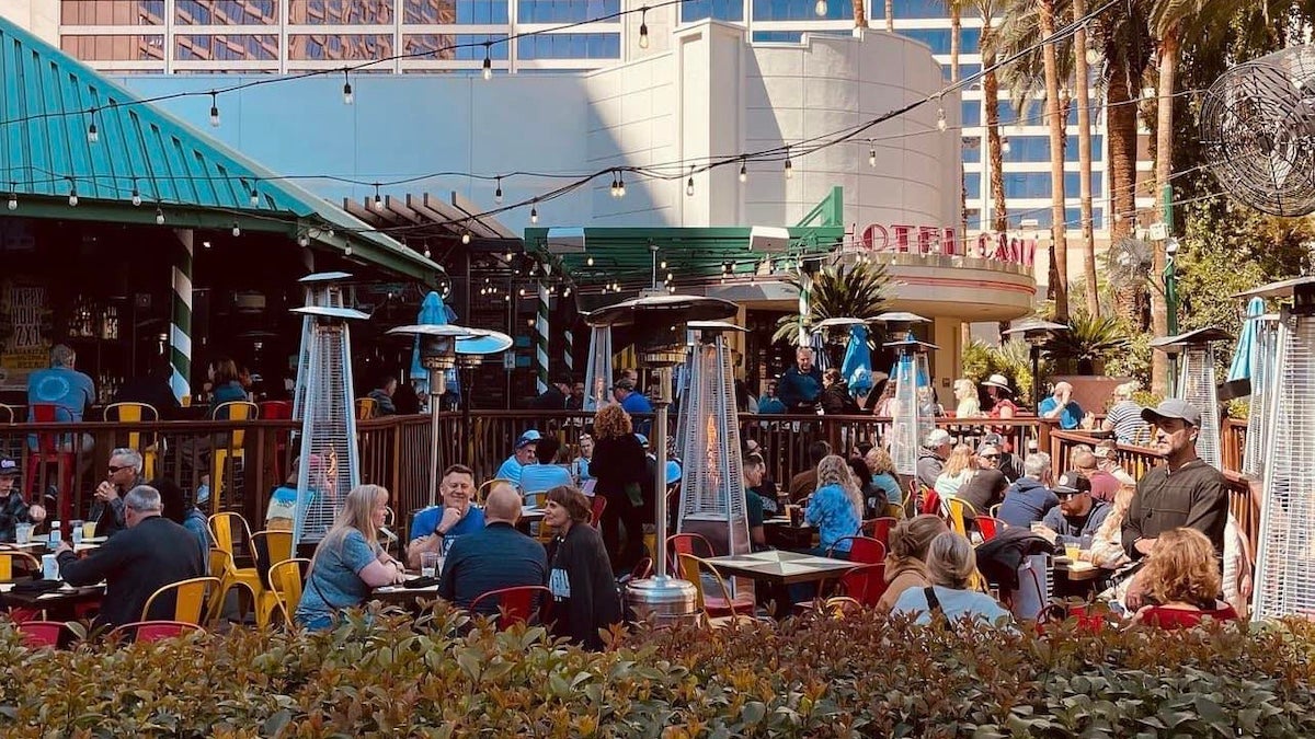 Outdoor patio dining scene at Carlos’n Charlie’s in Las Vegas, Nevada, with numerous people enjoying meals and drinks under string lights and surrounded by tall outdoor heaters, set against the backdrop of palm trees and high-rise buildings.