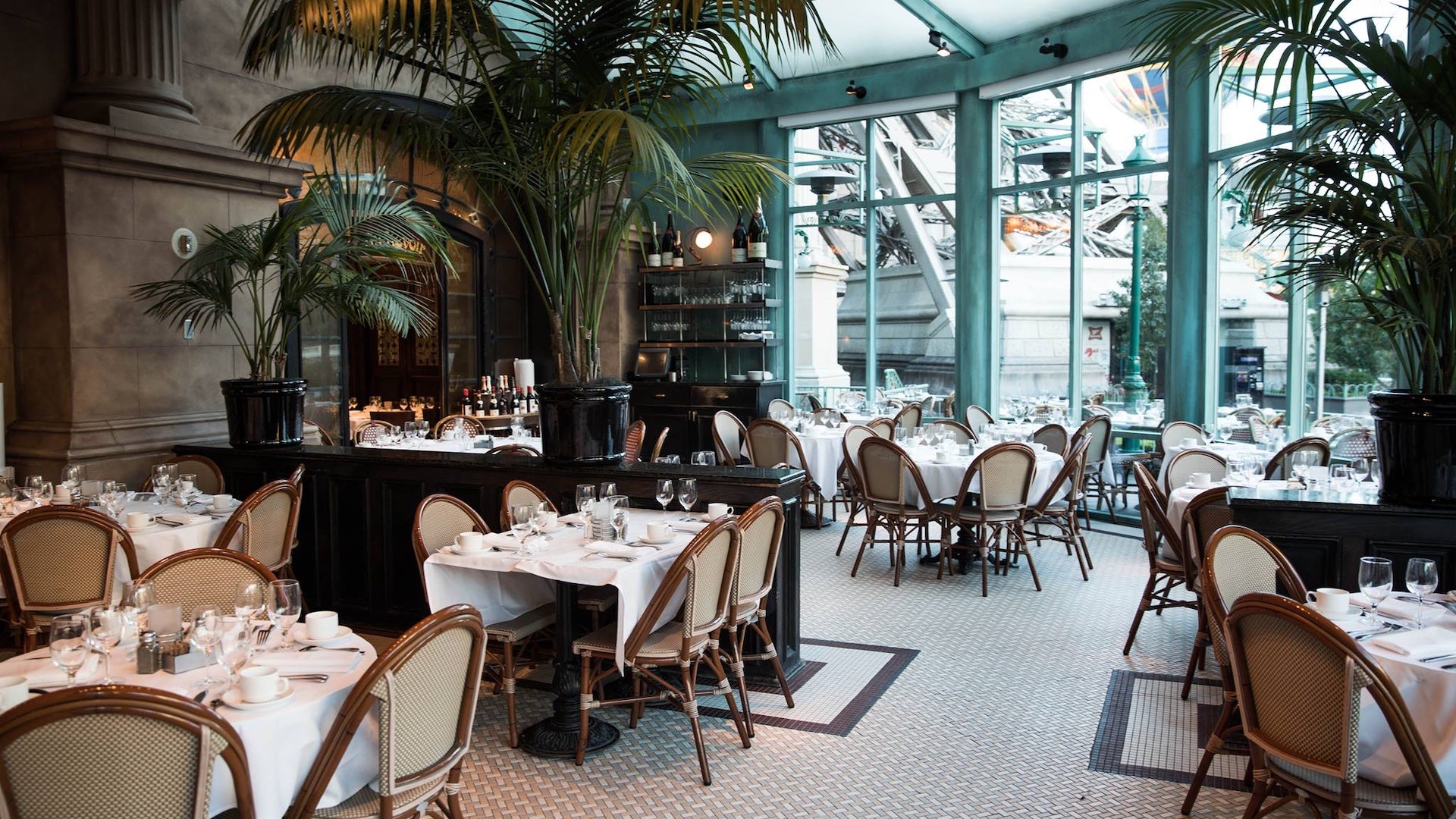 Elegant indoor dining area at Mon Ami Gabi in Las Vegas, featuring French bistro-style tables with white tablecloths, rattan chairs, tall potted palms, and large windows offering views of the Paris Las Vegas hotel exterior.