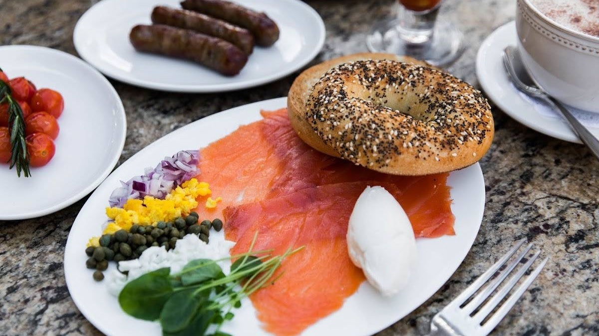 Plated lox and bagel breakfast at Mon Ami Gabi in Las Vegas, with an everything bagel, smoked salmon, cream cheese, chopped red onion, capers, boiled egg, microgreens, and a side of cherry tomatoes and sausages.