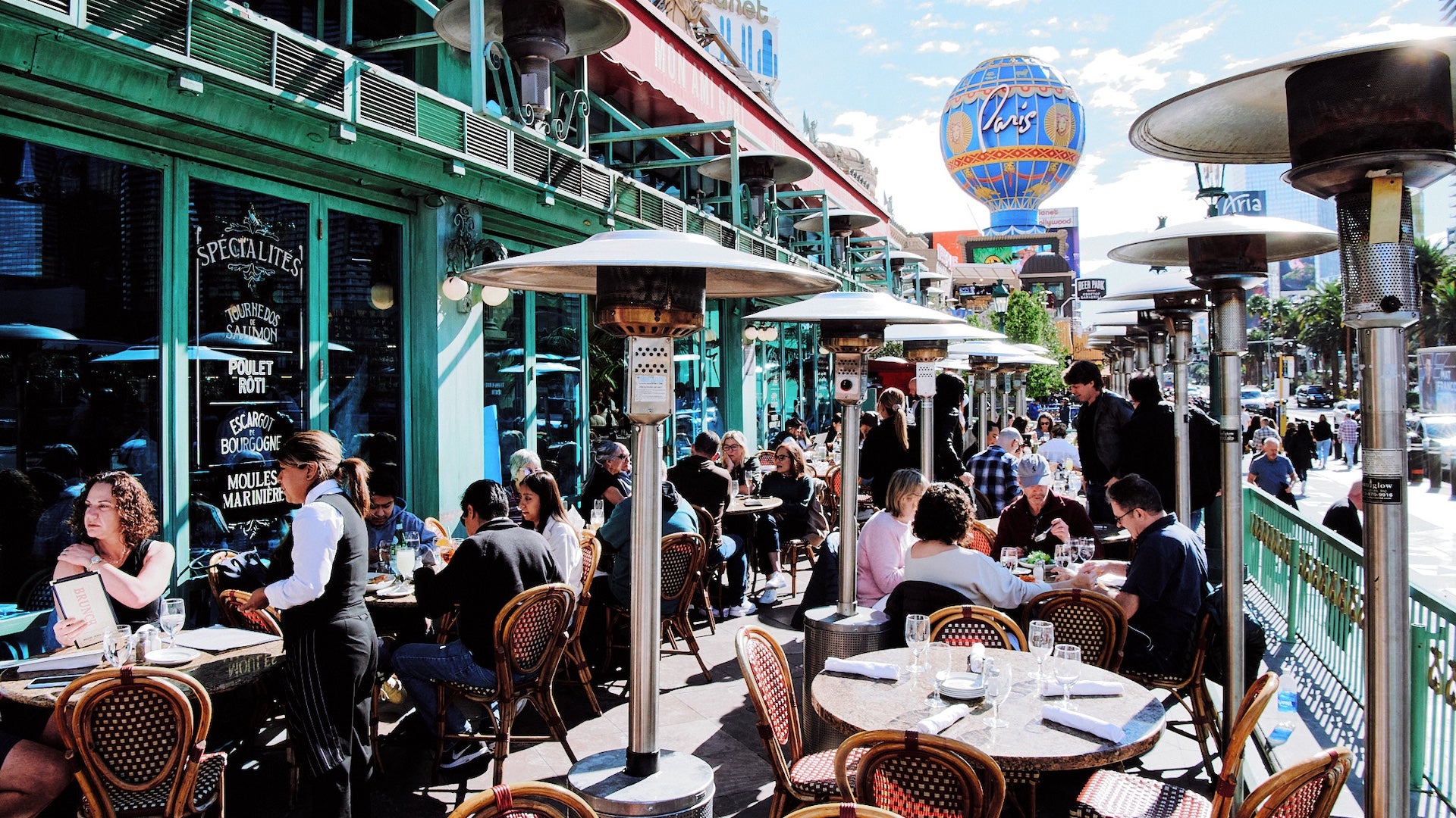 Bustling outdoor patio of Mon Ami Gabi on the Las Vegas Strip, filled with diners enjoying meals under heat lamps and shaded tables, with the Eiffel Tower replica and Paris Las Vegas balloon sign visible in the background.