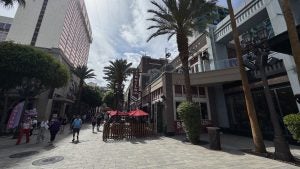 Pedestrian street scene at The LINQ Promenade in Las Vegas, featuring palm trees, outdoor diners under red umbrellas at Virgil’s Real BBQ, and surrounding shops and high-rise buildings under a partly cloudy sky.